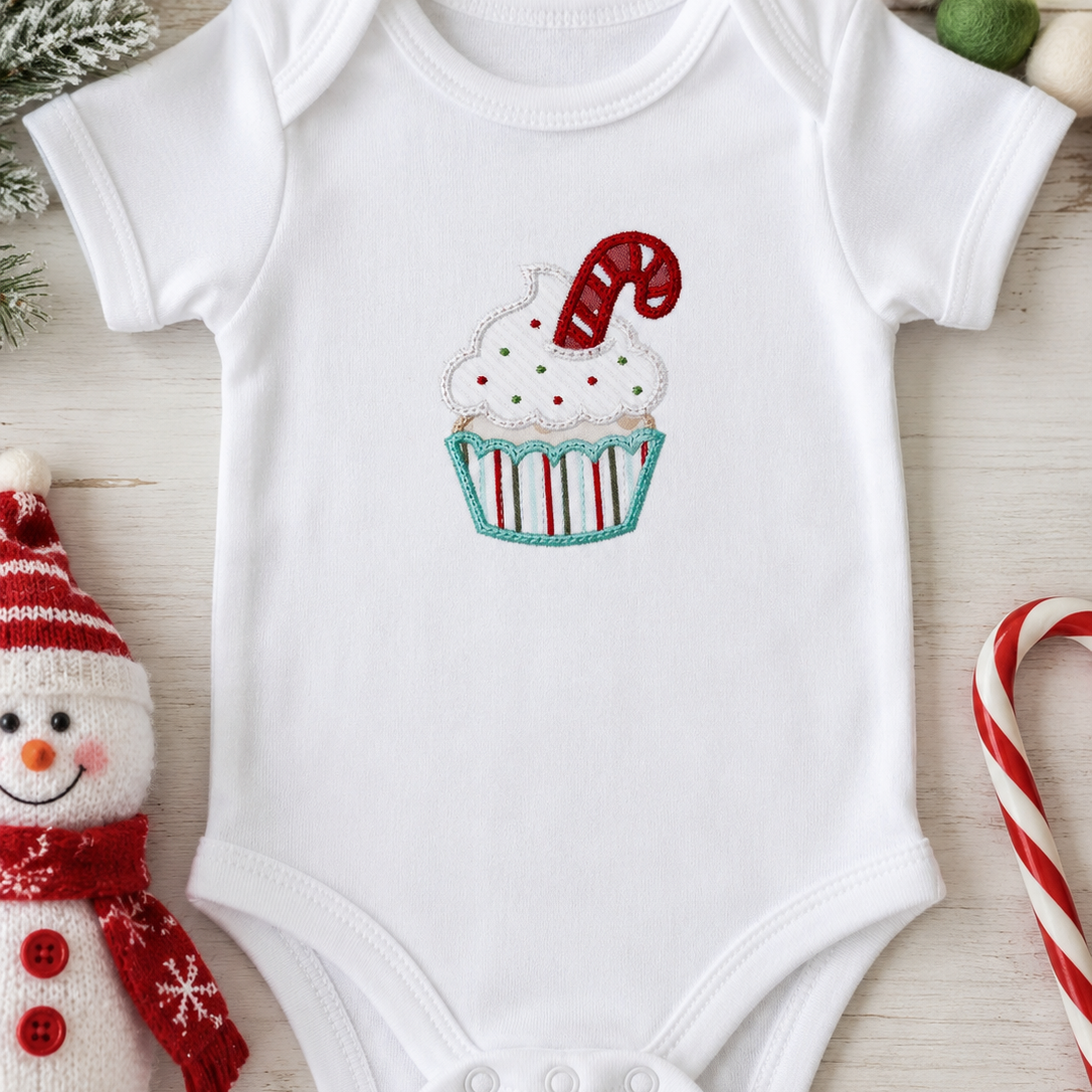 A white short-sleeve baby bodysuit laid flat on a light wood surface, featuring a cupcake graphic with white frosting, red and green sprinkles, and a red-and-white candy cane. The bodysuit is styled with holiday props including a snowman plush, candy canes, pinecones, and pom-poms.