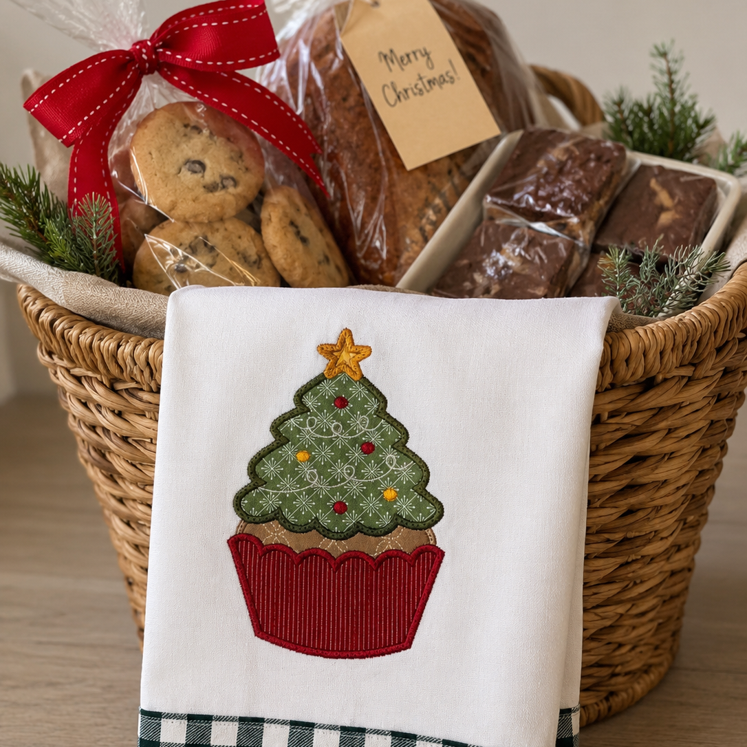 A holiday gift basket filled with cookies, chocolate bars, and wrapped treats, with a white tea towel draped over the front. The towel features an embroidered cupcake Christmas tree with a red gingham bottom border.