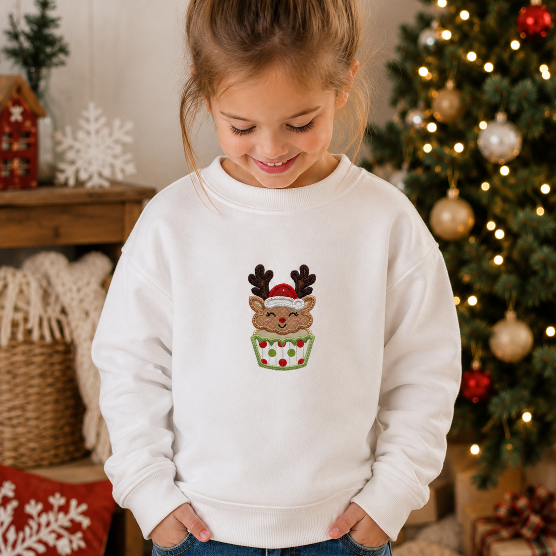 A young girl with her hair in a bun is wearing a white crewneck sweatshirt featuring a festive graphic of a reindeer wearing a Santa hat inside a green cupcake with red polka dots. The background shows a cozy Christmas setting with a decorated tree and holiday decorations.