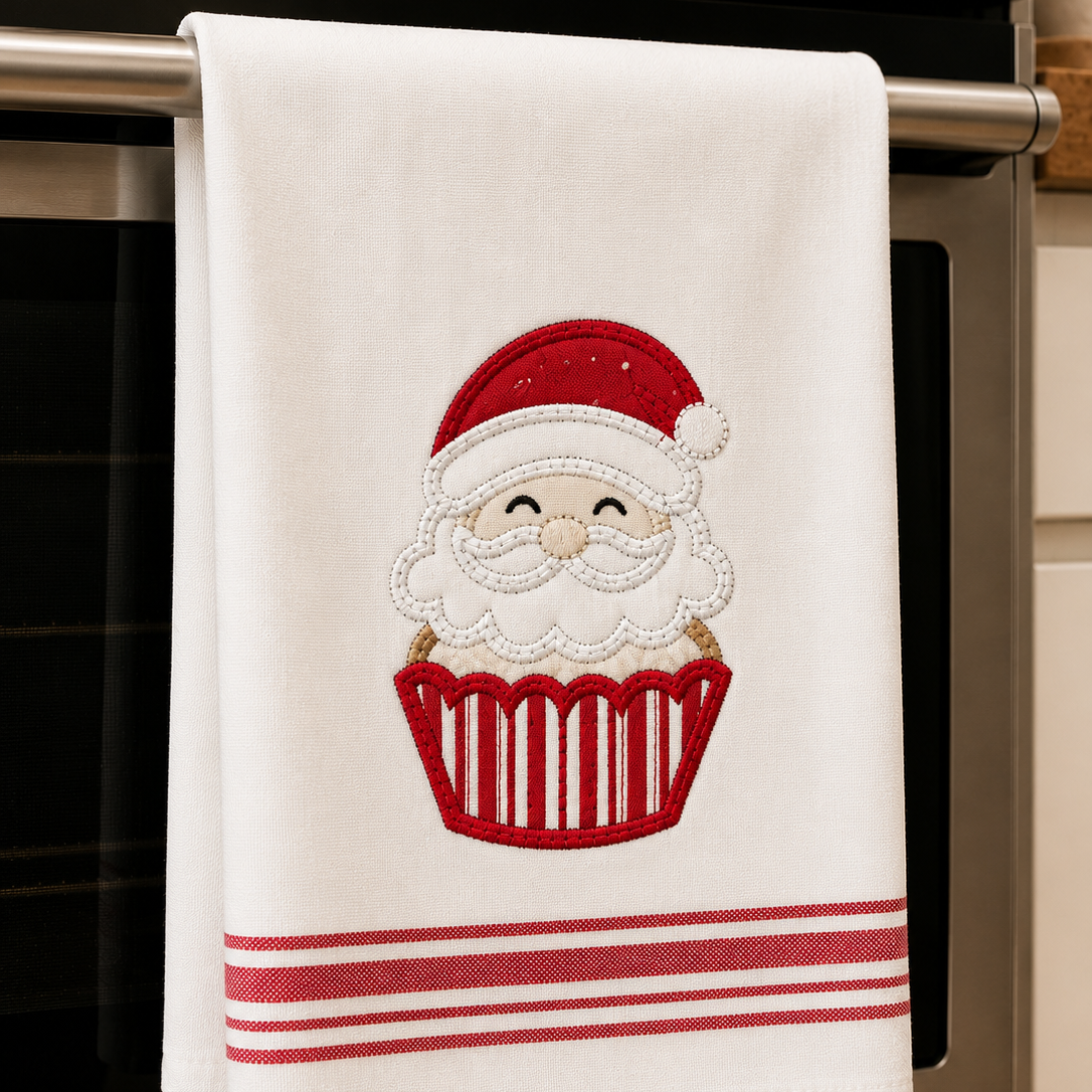 A white kitchen towel hanging from a stainless steel oven handle, featuring an embroidered Santa Claus face inside a red cupcake with red stripes at the bottom.