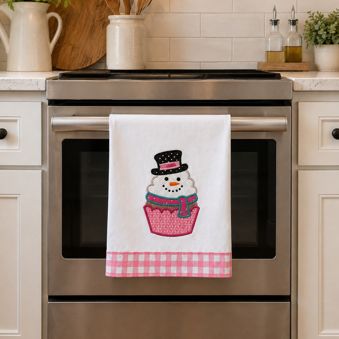 A white kitchen towel hanging on a stainless steel oven handle, featuring a cupcake with a snowman wearing a black top hat and a pink gingham bottom border. The scene is set in a bright, modern kitchen with white cabinets and a tiled backsplash.