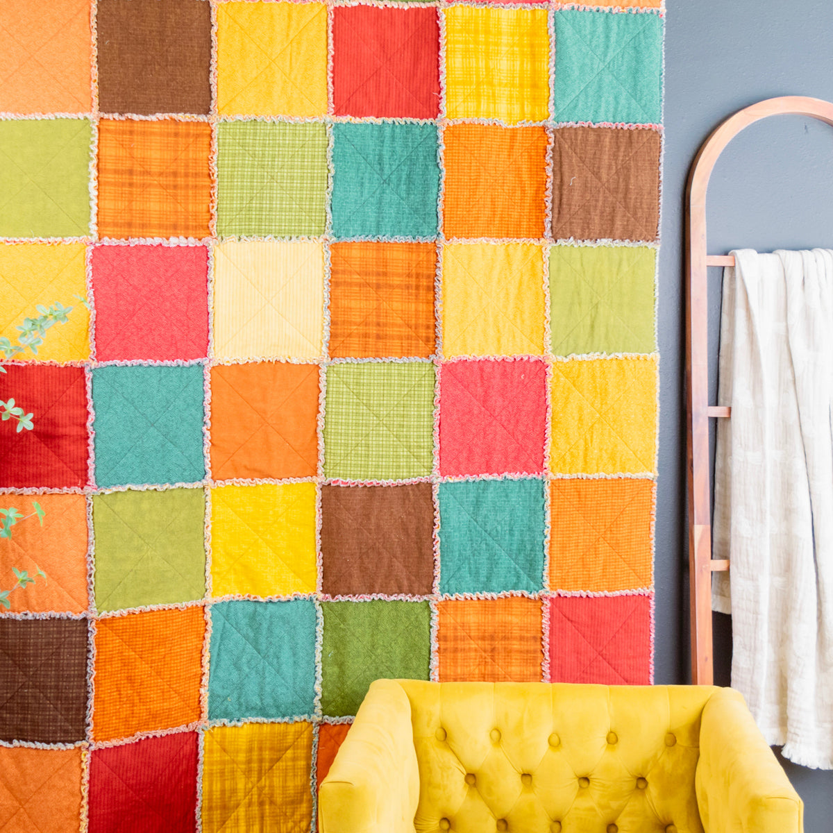 A colorful patchwork rag quilt hanging on a gray wall in a styled room, with a yellow tufted armchair at the bottom, a wooden towel ladder with a white towel on the right, and a green plant on the left.