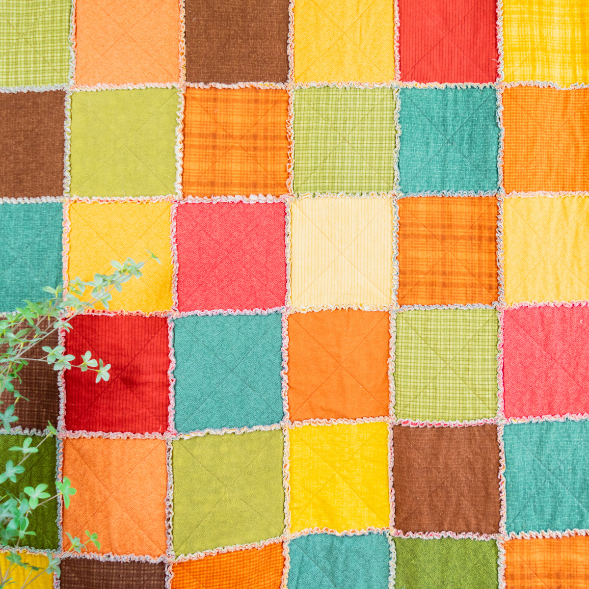 A colorful patchwork quilt hanging on a dark gray wall, featuring a grid of square fabric panels in orange, yellow, green, teal, red, brown, and cream, with a small green plant in the lower left corner.