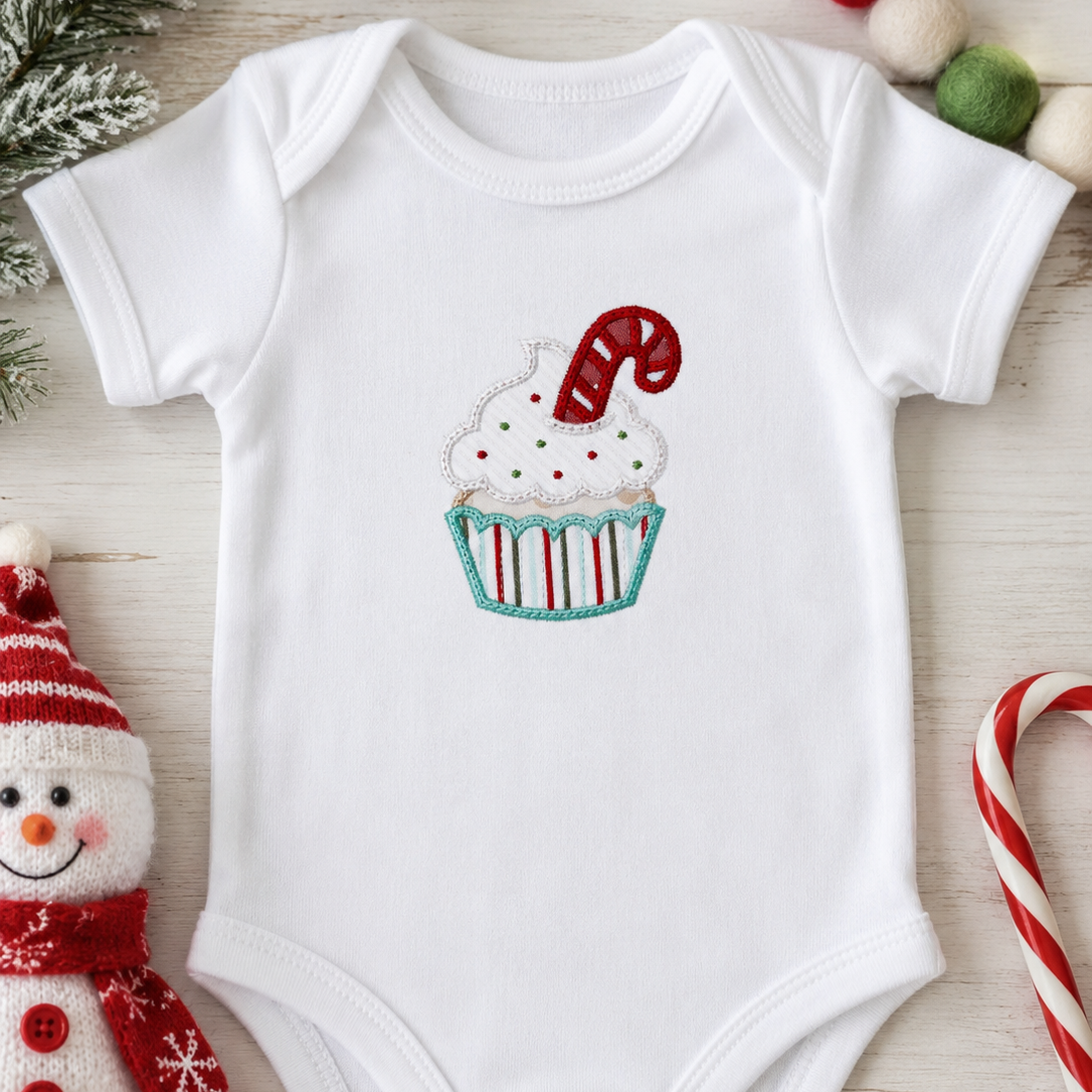 A white short-sleeve baby bodysuit laid flat on a light wood surface, featuring a cupcake graphic with white frosting, red and green sprinkles, and a red-and-white candy cane. The bodysuit is styled with holiday props including a snowman plush, candy canes, pinecones, and pom-poms.