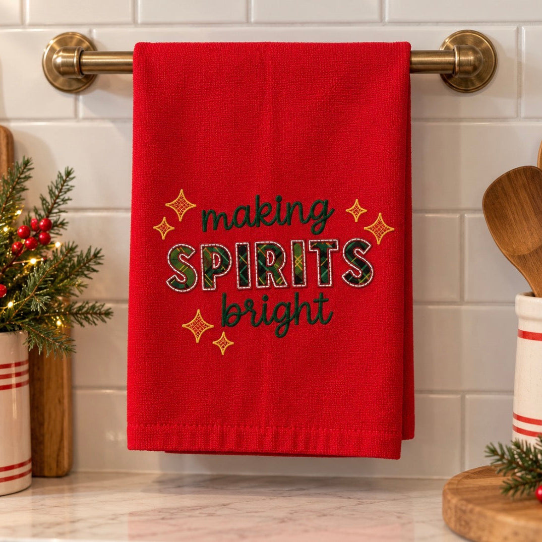 A red kitchen towel hanging on a brass towel bar, featuring the embroidered text 'making SPIRITS bright' with gold star accents, set against a white subway tile backsplash.