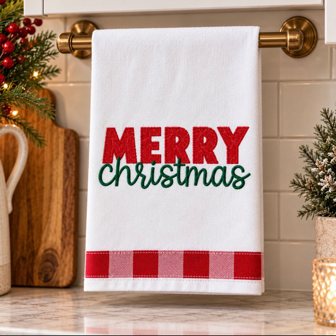 A white kitchen towel hanging on a brass towel rack, featuring the text 'MERRY Christmas' in red and green, with a red and white gingham stripe at the bottom. The towel is displayed in a decorated kitchen setting with holiday greenery, a cutting board, and a marble countertop.