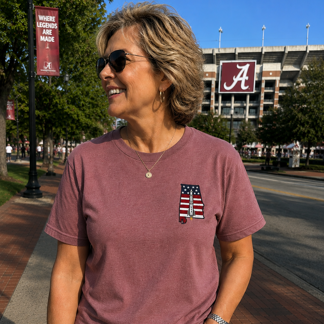 Woman wearing a maroon t-shirt with a logo, standing on a sidewalk with a stadium in the background.