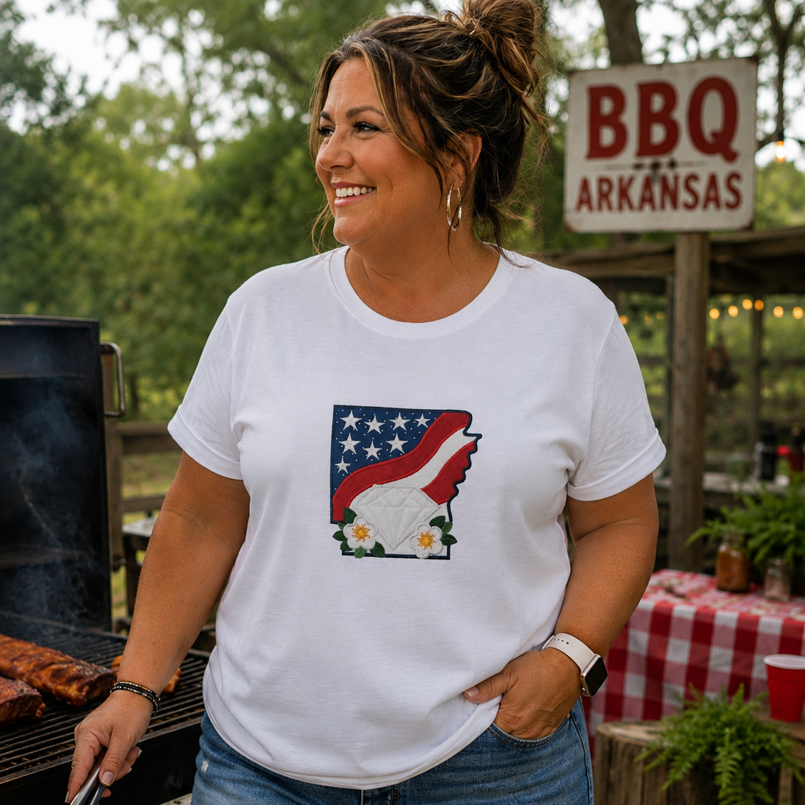 Woman wearing a white t-shirt with a graphic design, standing outdoors near a grill and 'BBQ Arkansas' sign.
