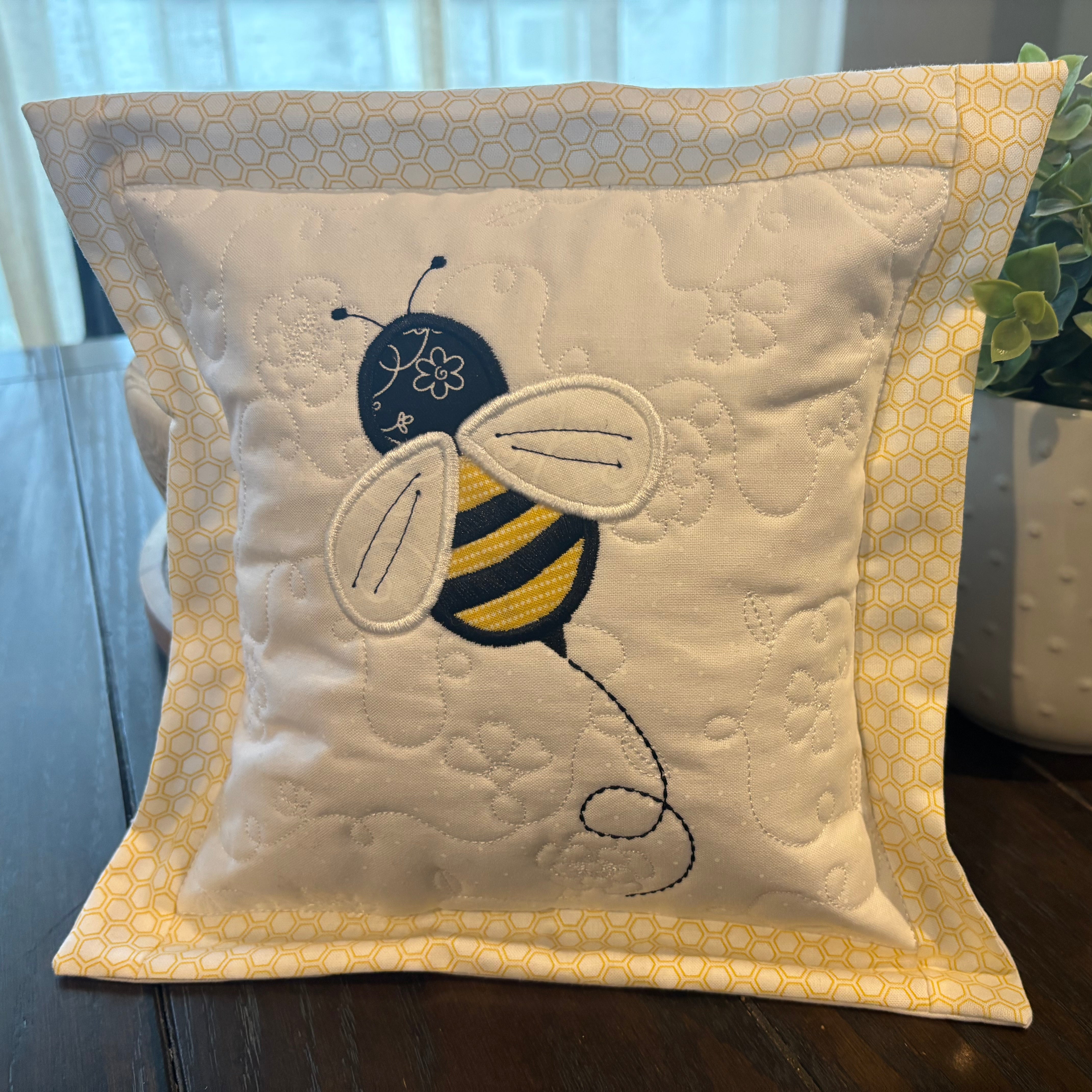 Decorative pillow with a bee design on a wooden surface next to a plant.