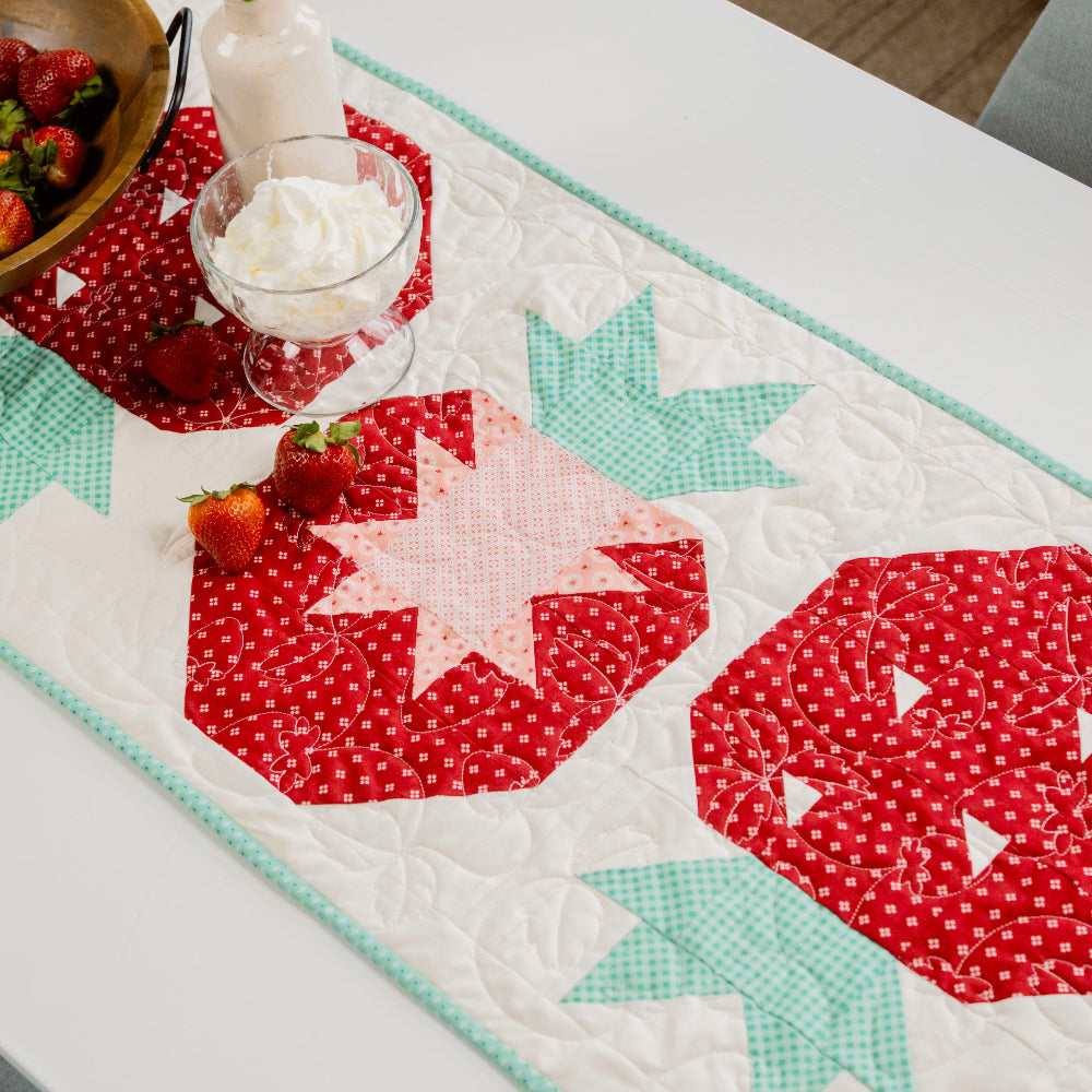 Table setting with a strawberry-themed quilted table runner, strawberries, and whipped cream.