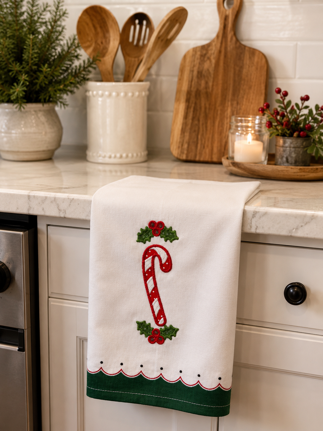 A white kitchen towel with a red and green embroidered candy cane and holly design, hanging over a white kitchen counter with kitchen utensils and plants in the background.