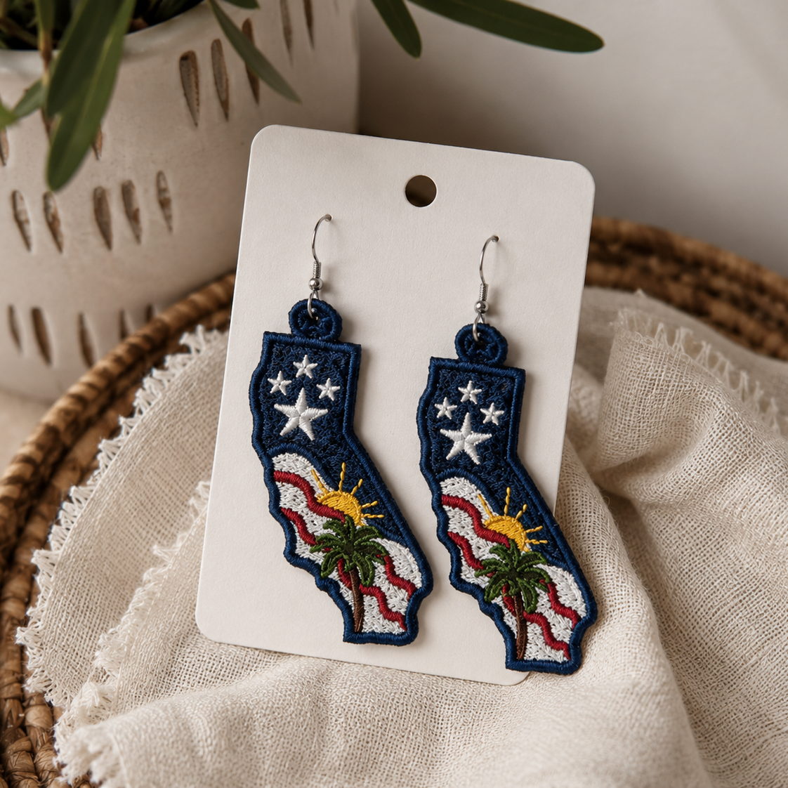 Pair of earrings on a card with a decorative design, placed on a textured surface with a plant in the background.
