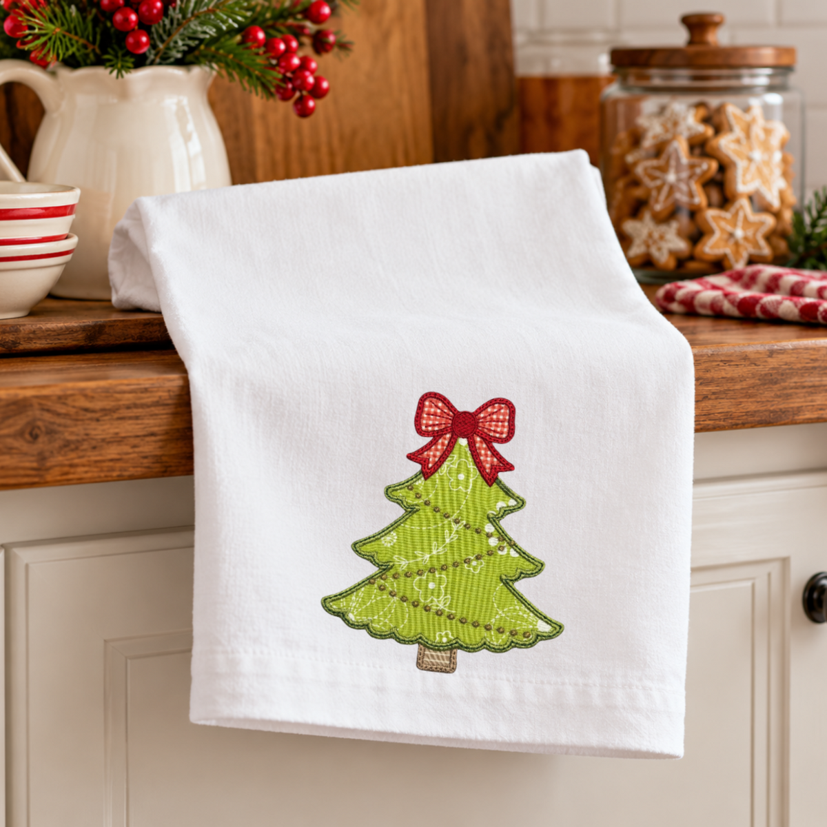A white kitchen towel draped over a wooden countertop, featuring an embroidered green Christmas tree with a red bow on the front.