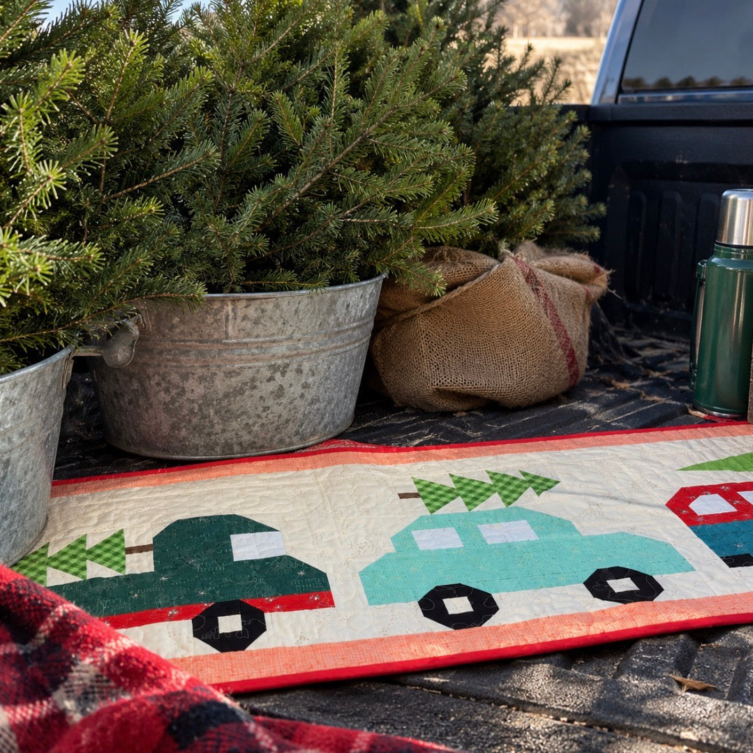A rustic quilted table runner featuring cartoon-style Christmas trucks carrying green trees, styled in a pickup truck bed with potted Christmas trees and burlap.