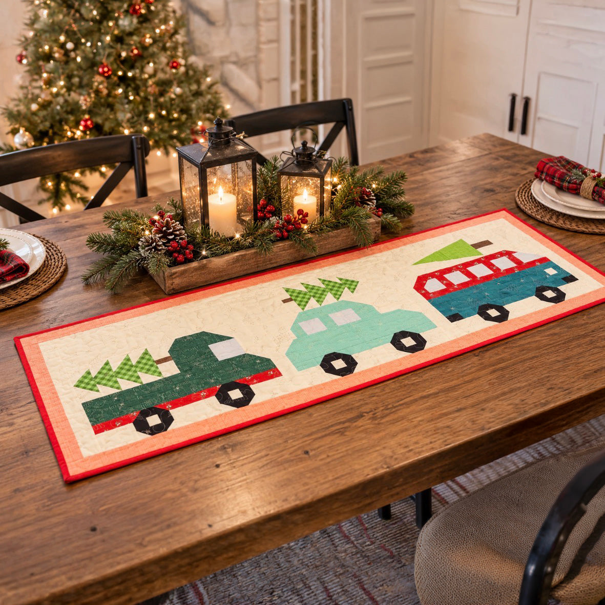 A festive holiday table runner featuring three colorful Christmas trucks carrying tree decorations, styled on a wooden dining table with candles, lanterns, and a decorated Christmas tree in the background.