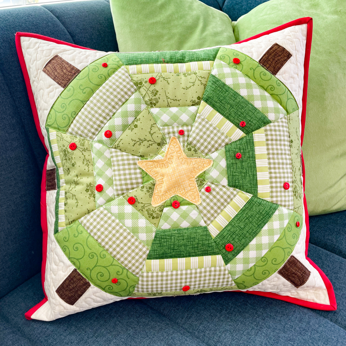 A square quilted throw pillow with a green and white gingham and floral patchwork design, featuring a central tan star and red button accents, resting on a blue couch with green pillows in the background.