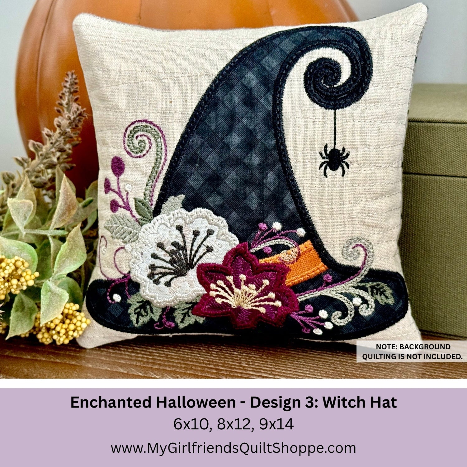 A square decorative pillow shown front-facing on a tabletop with autumn props. The pillow features a large embroidered witch hat design in black and navy with floral accents in burgundy, white, purple, and orange.