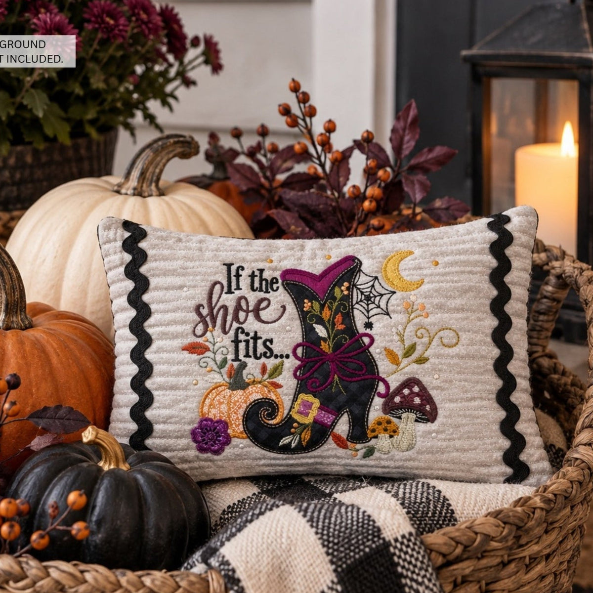 A decorative square throw pillow displayed in a woven basket with pumpkins, fall foliage, and a candle. The pillow has a cream fabric background with black scalloped trim and an embroidered Halloween design featuring a black boot, purple bow, mushrooms, spiderwebs, and the text 'If the shoe fits.