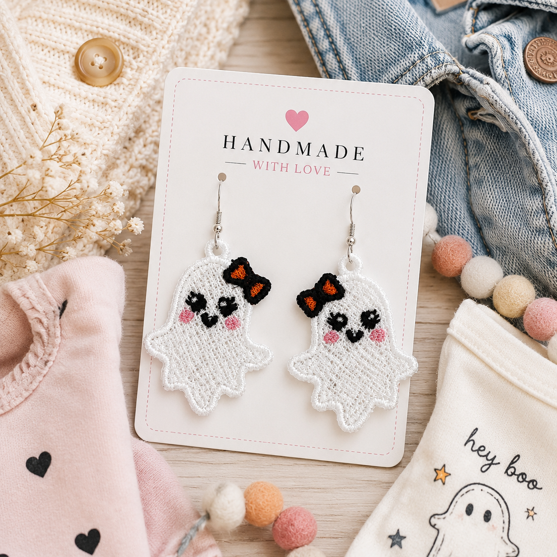 A pair of handmade embroidered ghost dangle earrings displayed on a white card labeled 'HANDMADE WITH LOVE'. The card is styled in a flat-lay arrangement with baby clothing, a denim jacket, and pastel pom-pom beads on a wooden surface.