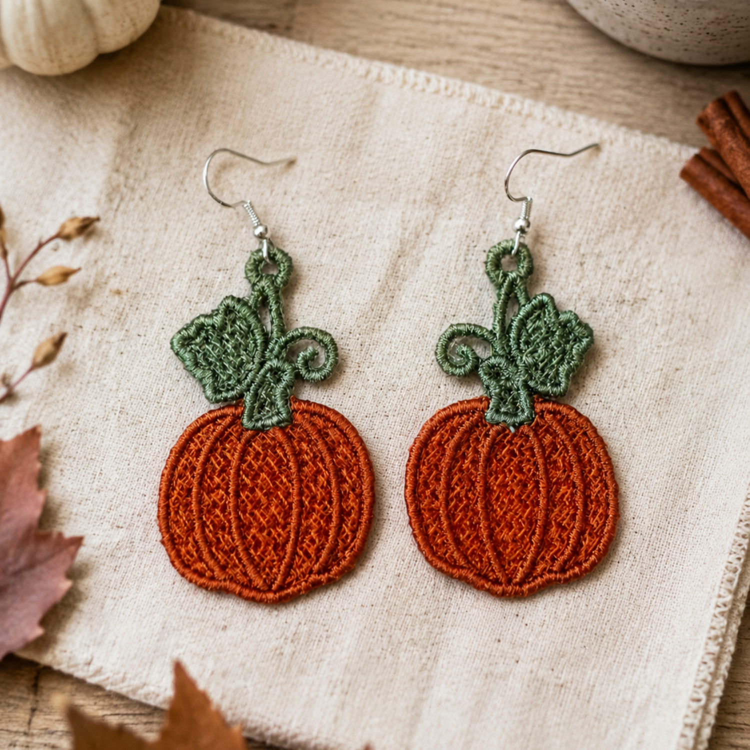 A pair of handmade embroidered pumpkin earrings with green stems and silver fishhook wires, displayed on a textured cream linen cloth with autumn props like a small white pumpkin, cinnamon sticks, and leaves.