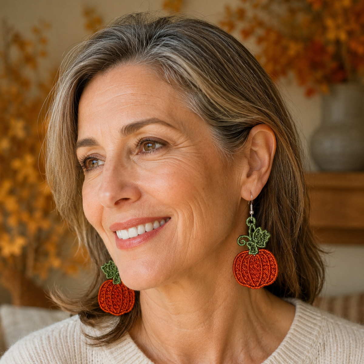 A smiling woman with gray-brown hair wearing large orange pumpkin-shaped drop earrings with green leaf details, photographed in a warm indoor setting with autumn foliage in the background.