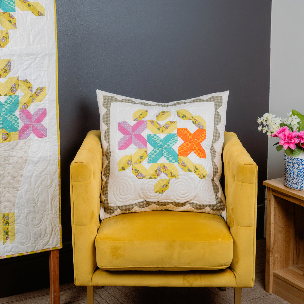 Yellow armchair with a quilted pillow featuring colorful floral patterns, next to a quilt on a stand against a gray wall.