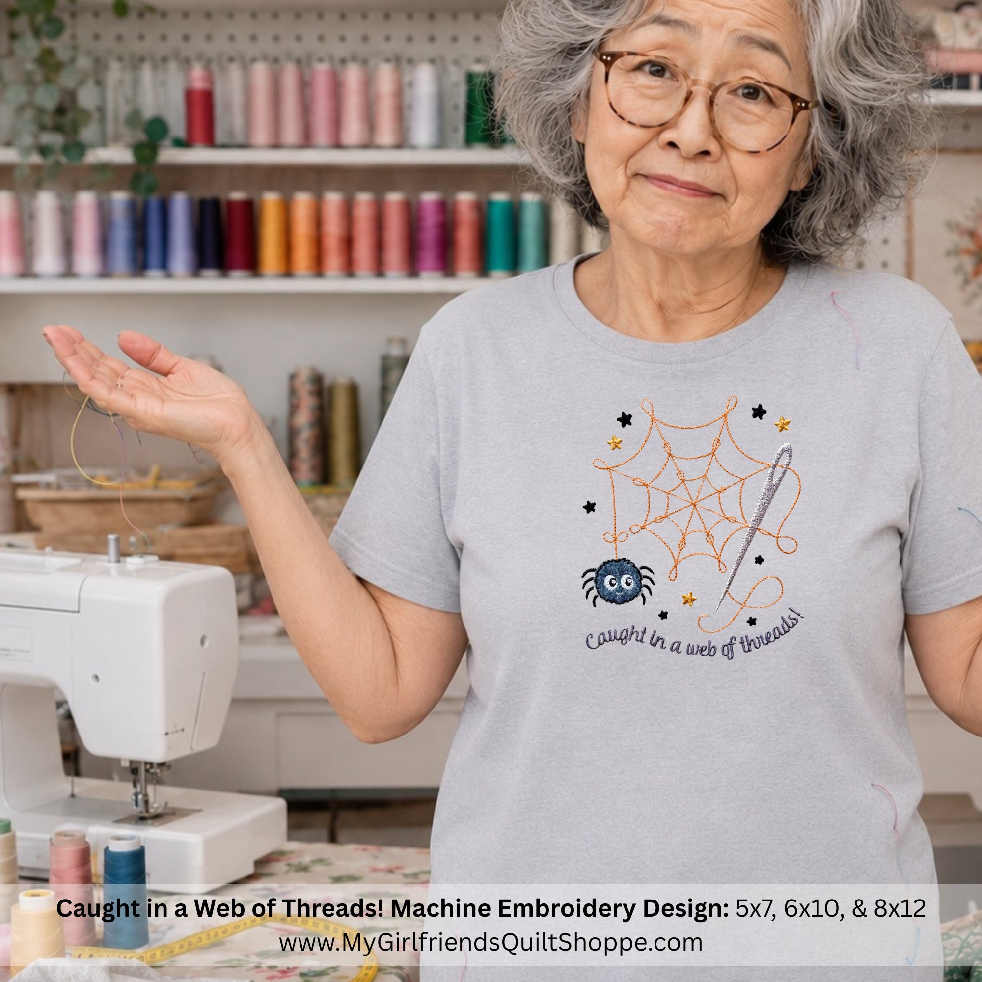 An older woman with glasses stands in a sewing room wearing a light gray t-shirt with an embroidered spiderweb and spider design. A white sewing machine is visible on the left, and shelves of colorful thread spools are in the background.