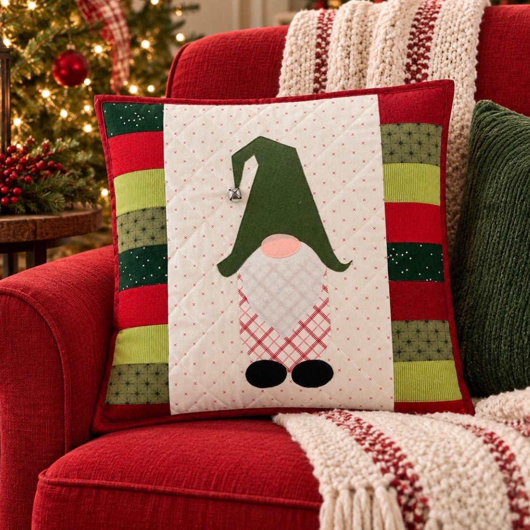 A square Christmas throw pillow with a gnomes face design, styled on a red armchair next to a green knit pillow and a cozy knit blanket, with a decorated Christmas tree in the background.