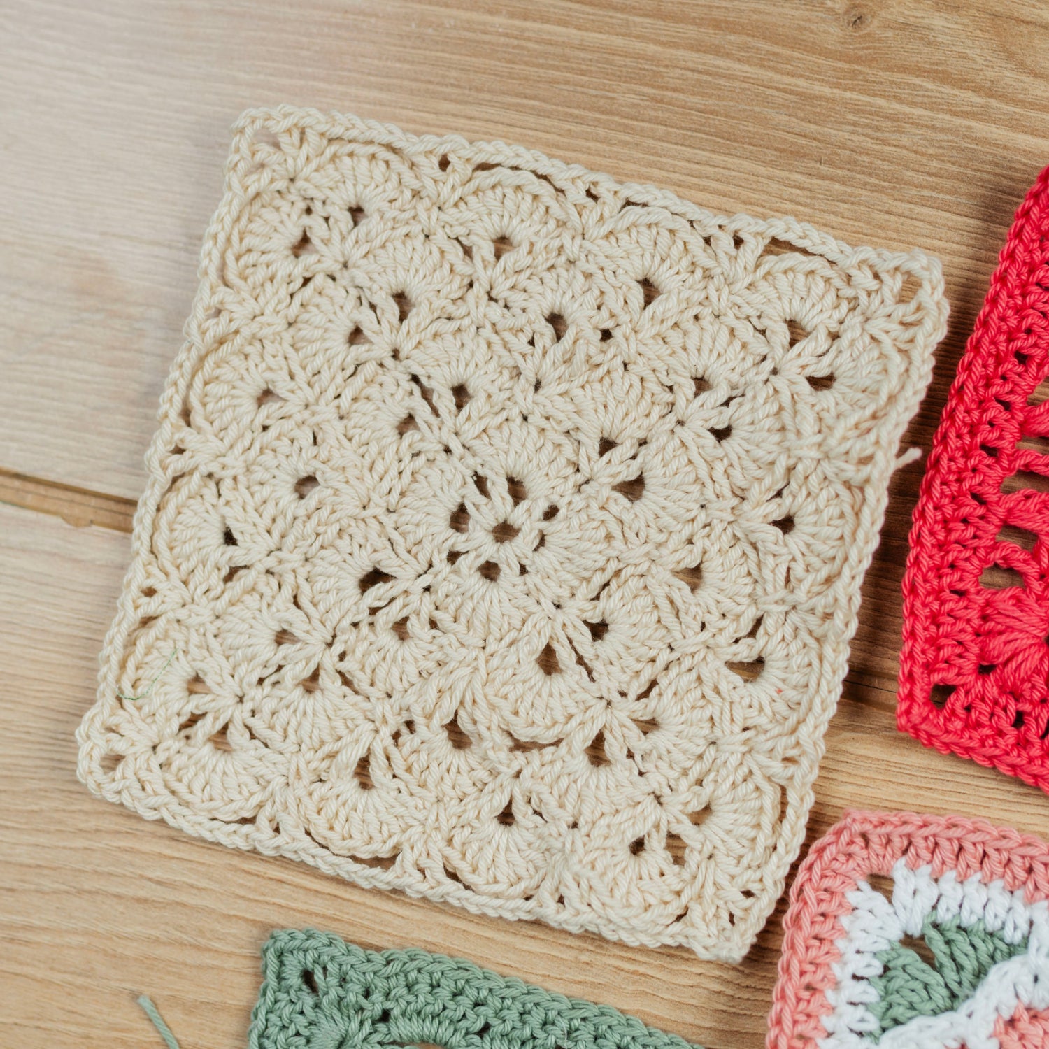 A close-up of a handmade crocheted granny square in a warm cream color, displayed on a light wood background next to other colorful crochet squares.