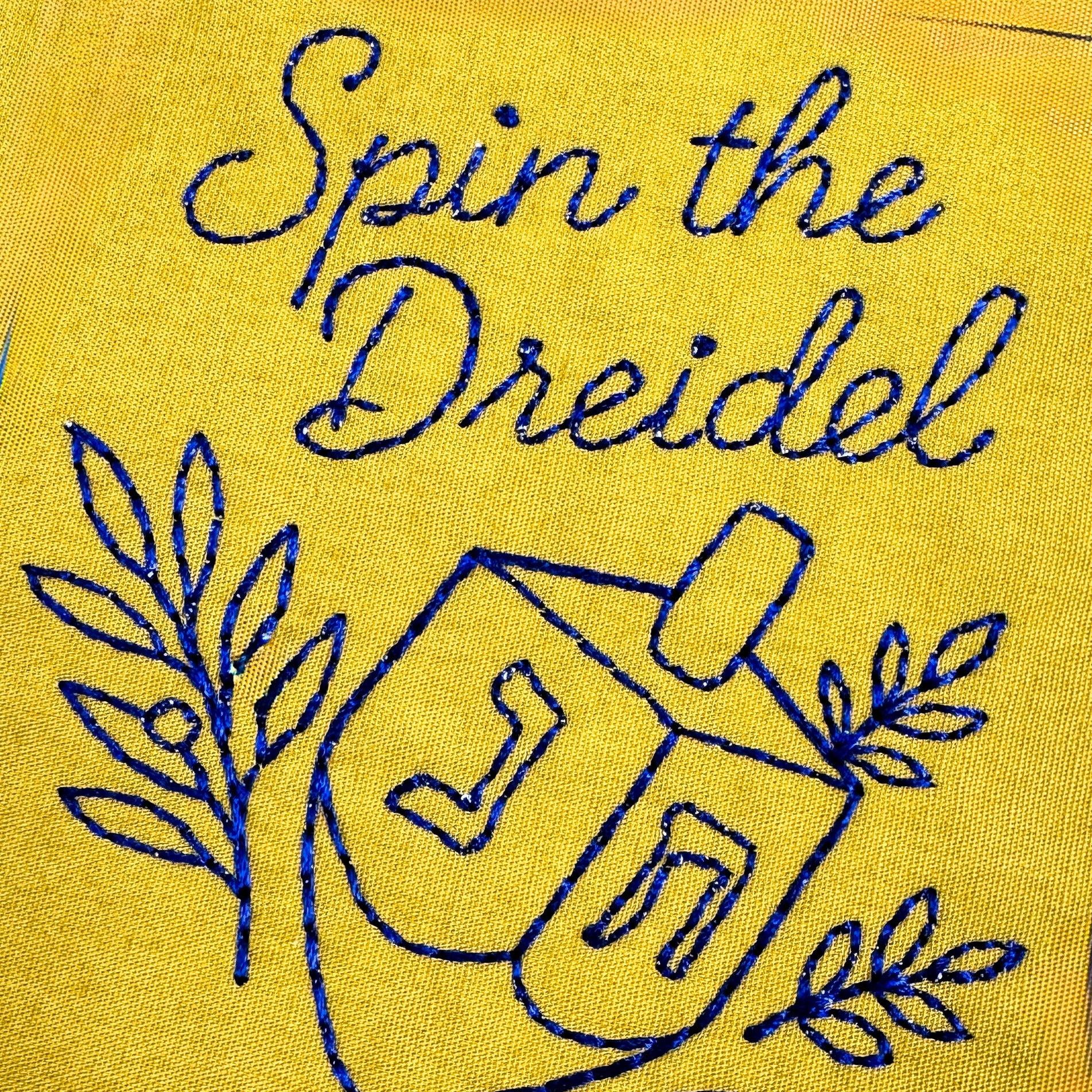 Yellow fabric with blue embroidered design of a dreidel and text 'Spin the Dreidel'.