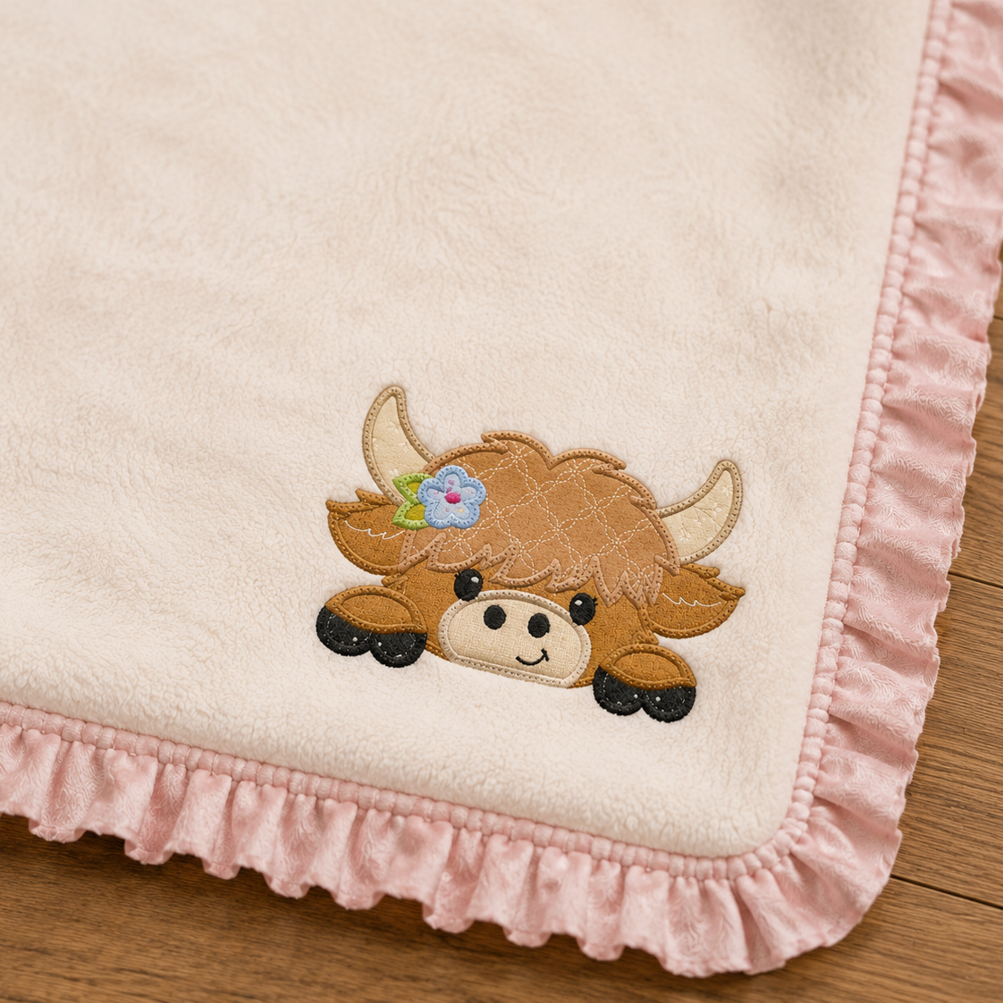 A close-up of a soft cream fleece blanket with a pink ruffled border, featuring an embroidered brown highland cow character with horns and a small flower on the corner.
