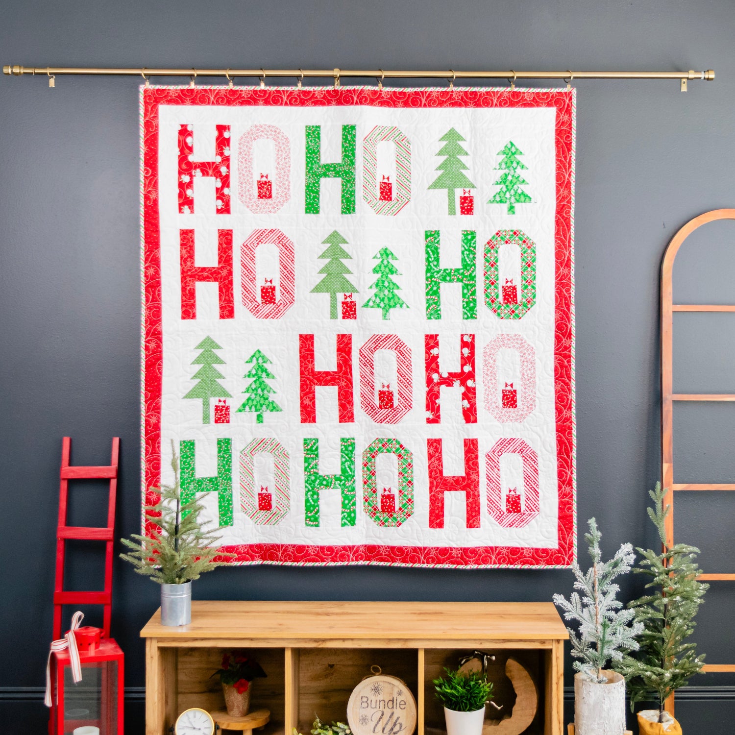 A Christmas-themed quilt hanging on a gold rod against a dark gray wall, featuring the word 'HO HO HO' in red and green with small tree and cabin motifs. The quilt is displayed in a styled room setting above a wooden console table with plants and decor.