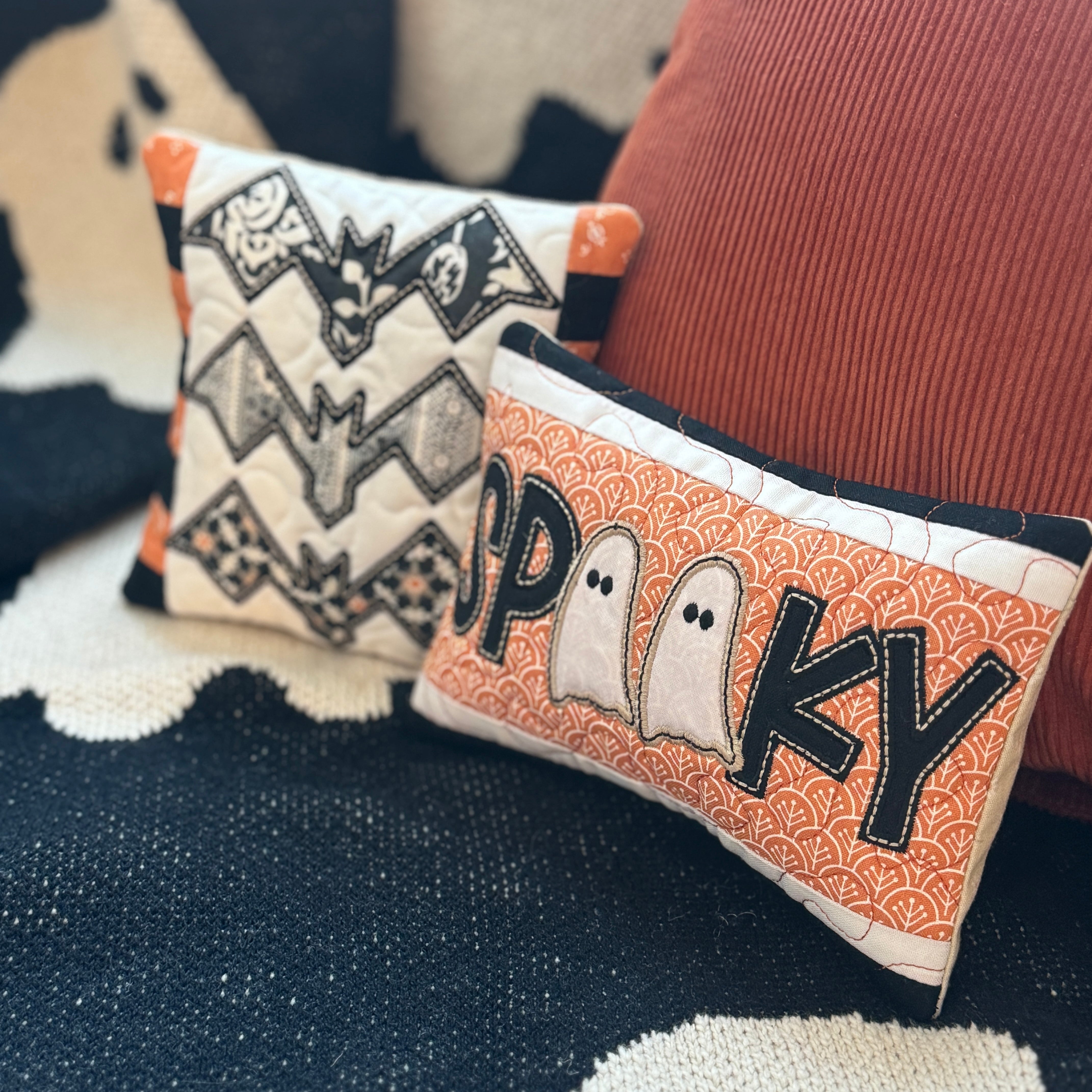 Two Halloween-themed pillows with bat and ghost designs on a textured fabric background.