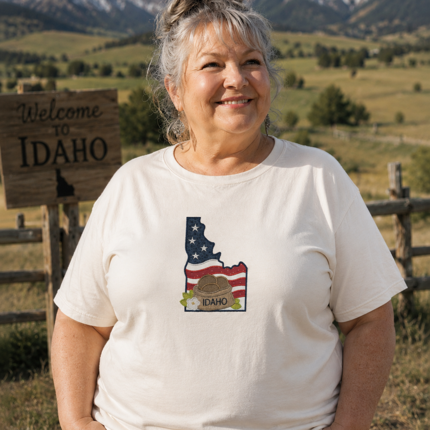 Woman wearing a white t-shirt with Idaho design, standing in front of a scenic landscape with mountains and a 'Welcome to Idaho' sign.