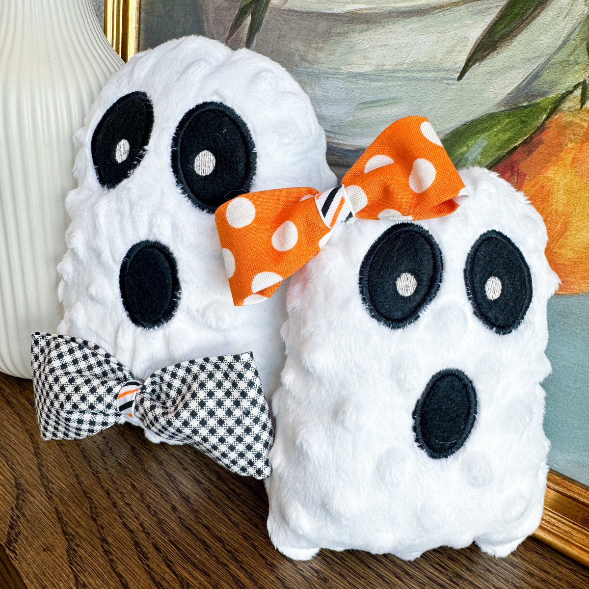 Two plush ghost toys with decorative bows on a wooden surface, with a painting of oranges in the background.