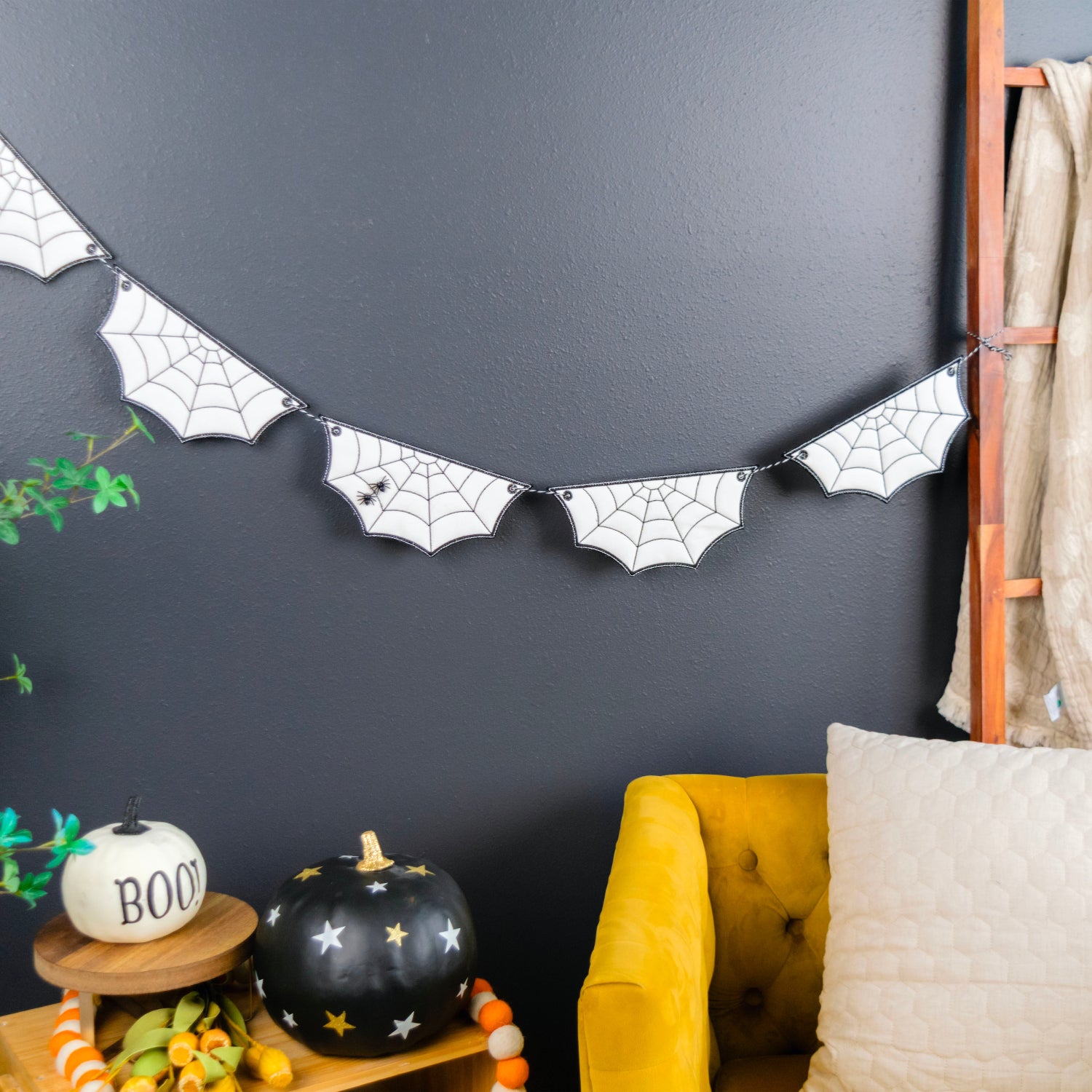 A Halloween-themed bat banner with black spiderweb designs hangs on a dark gray wall in a styled room setting with a yellow armchair, decorative pumpkins, and a plant.