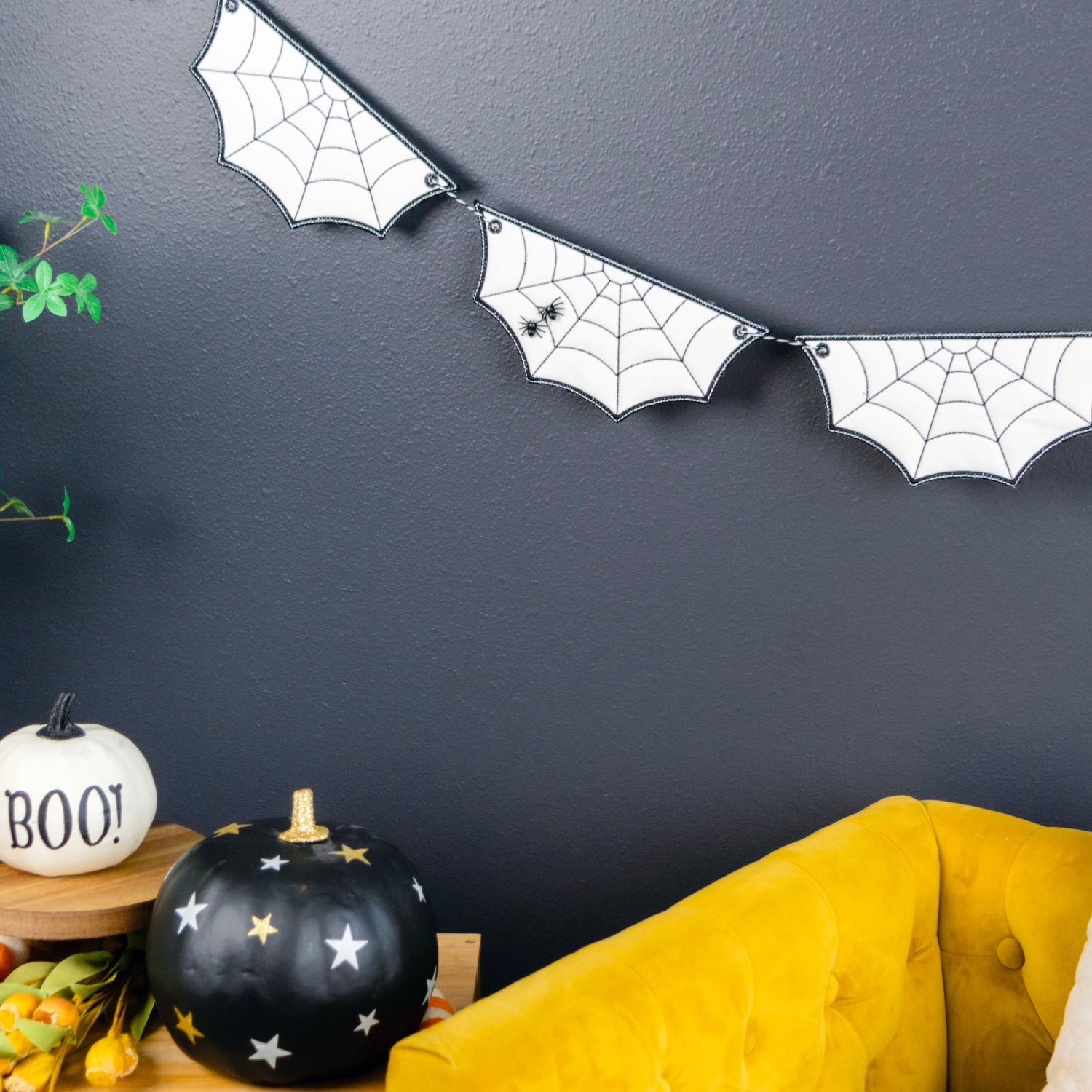 A Halloween-themed banner of white bat-shaped flags with black spiderweb patterns hanging on a dark gray wall above a yellow tufted sofa and a wooden shelf with decorative pumpkins.