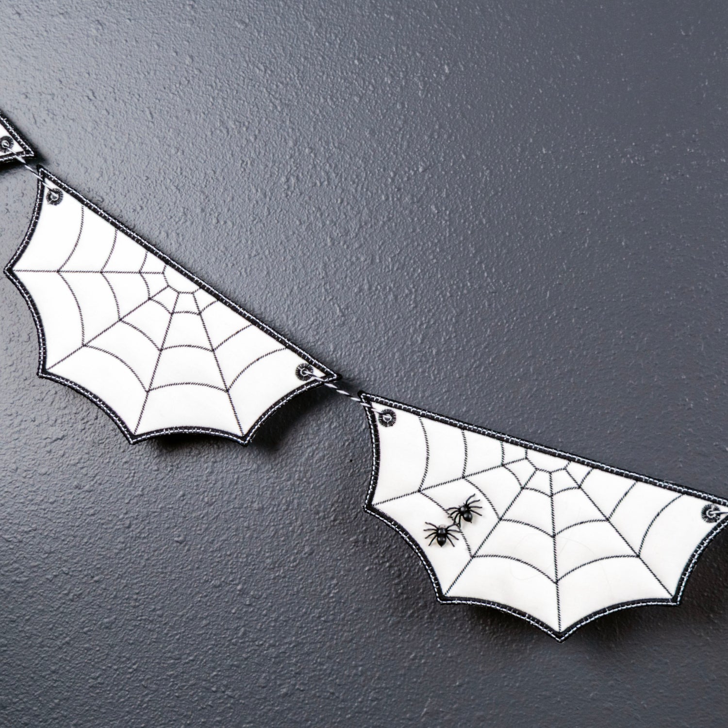 White bat-shaped banner flags with black spiderweb patterns and small black spider decorations, strung together against a dark textured wall with a small green plant branch in the corner.