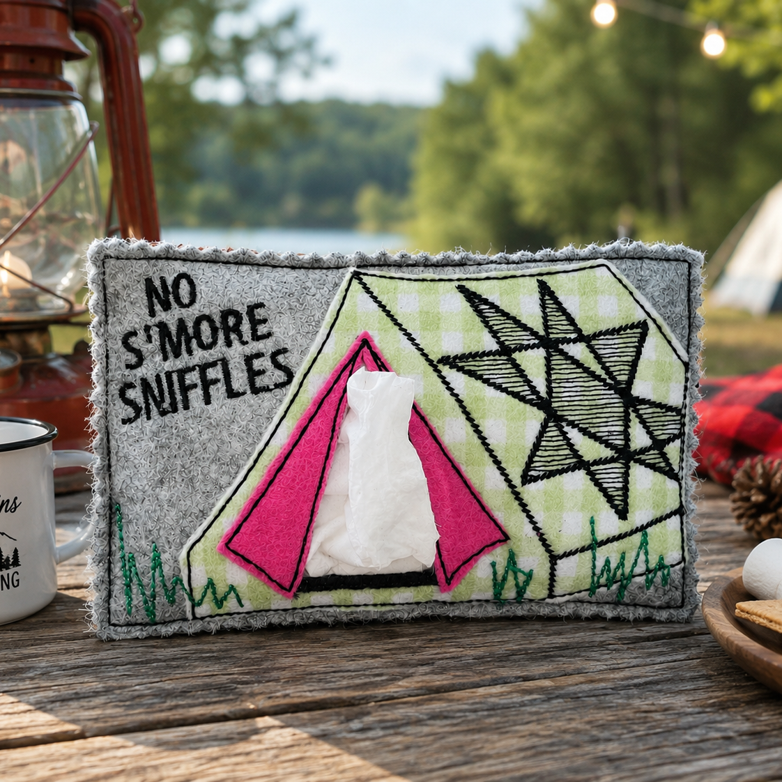 A felt camping-themed tissue box cover sits on a wooden table outdoors, with a lantern, mug, marshmallows, and campsite background visible. The cover has a gray border, a green-and-white plaid background, black embroidered text reading 'NO S'MORE SNIFFLES,' a pink tent motif, and a black geometric star design.