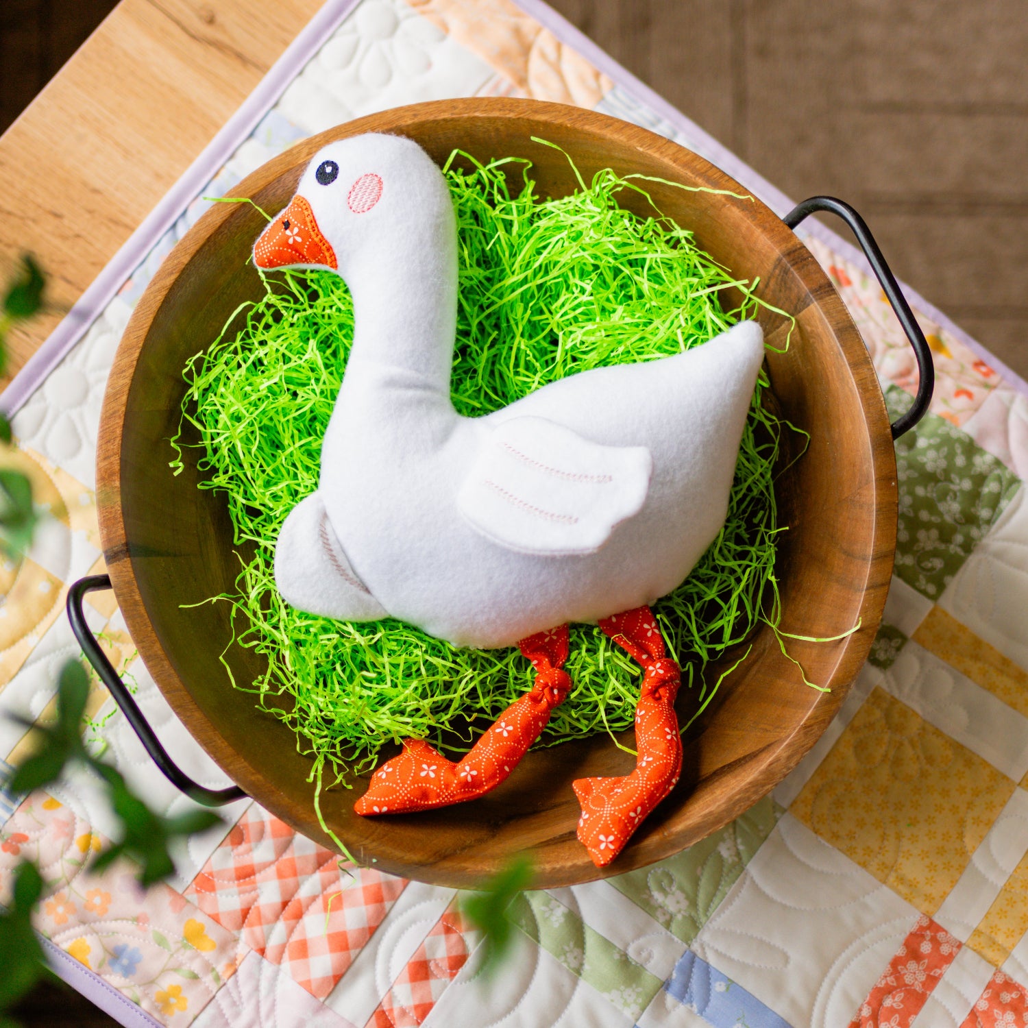 White goose figurine with orange feet and beak on green grass in a wooden bowl on a patterned tablecloth.