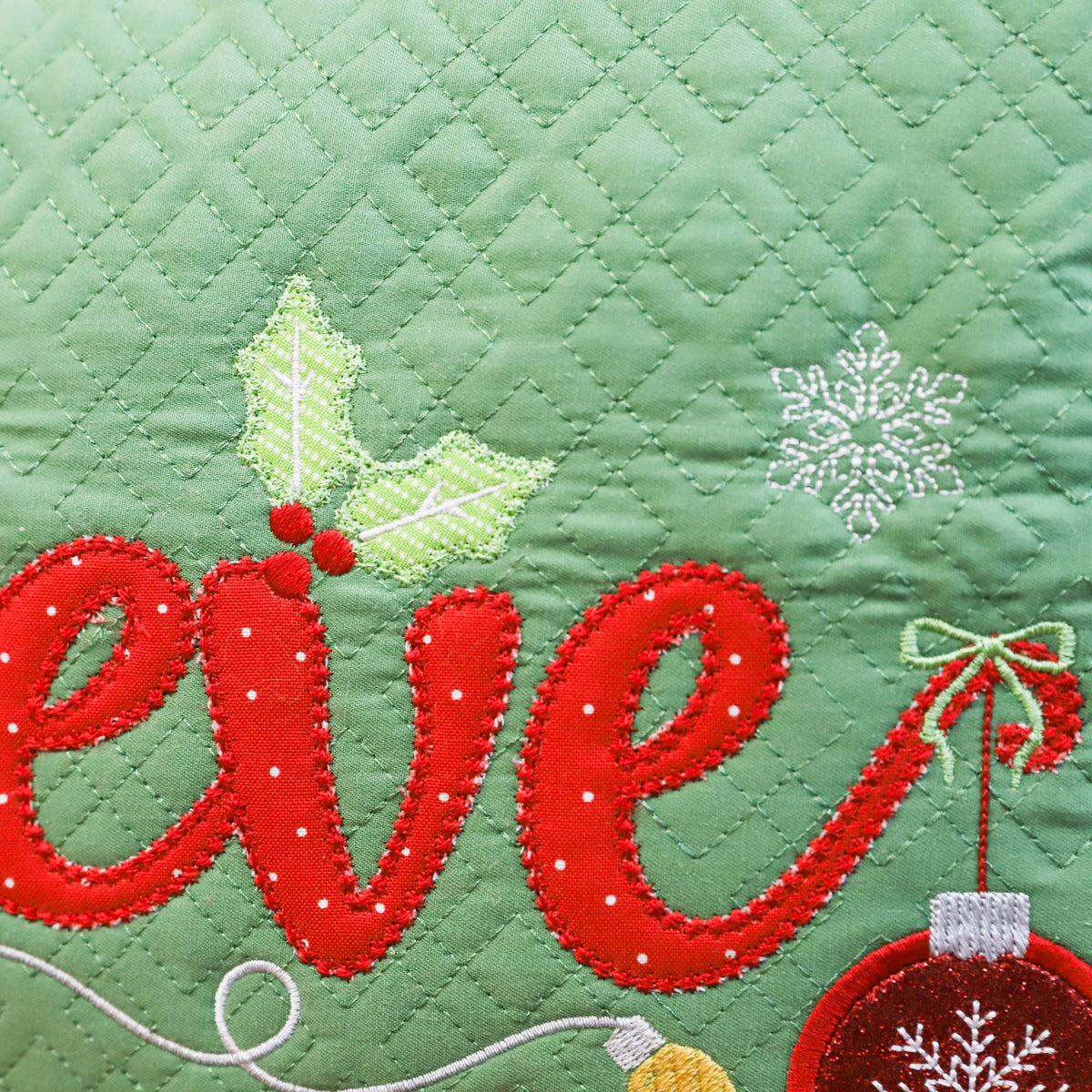 Close-up of a green quilted fabric with embroidered holiday lettering reading "eve" in red, white snowflakes, holly leaves, and a red ornament.