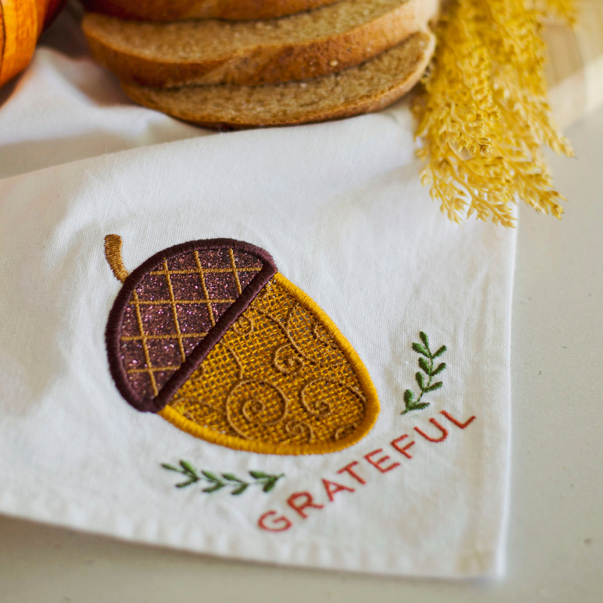 A white fabric napkin or kitchen towel with an embroidered acorn design and the word 'GRATEFUL' in red, styled with bread slices, a pumpkin, and dried yellow grass.