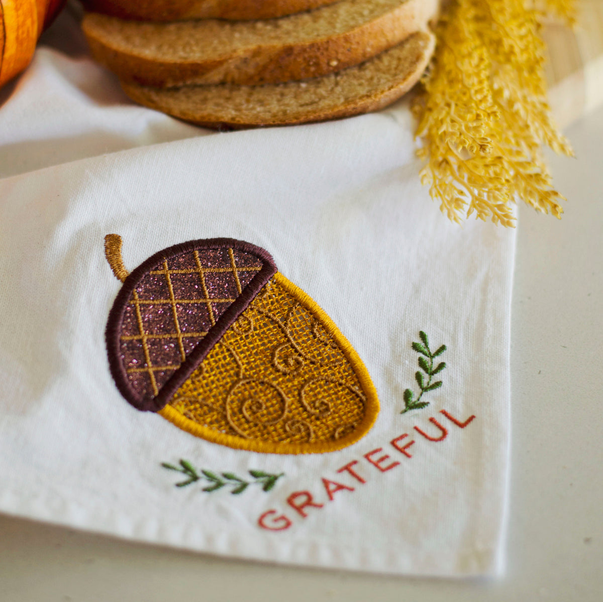 A white fabric napkin or kitchen towel with an embroidered acorn design and the word 'GRATEFUL' in red, styled with bread slices, a pumpkin, and dried yellow grass.