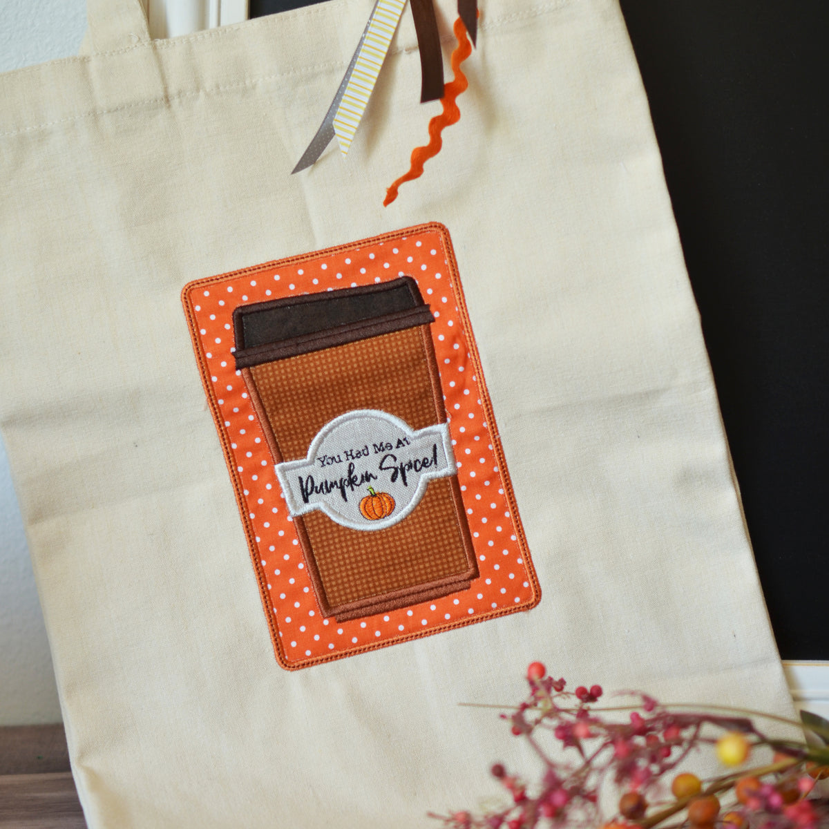 A cream canvas tote bag featuring a large applique patch of an orange coffee cup with white polka dots and a 'You Are So Warm Pumpkin Spice' sticker. The bag is styled with autumn-themed ribbons and dried flowers.