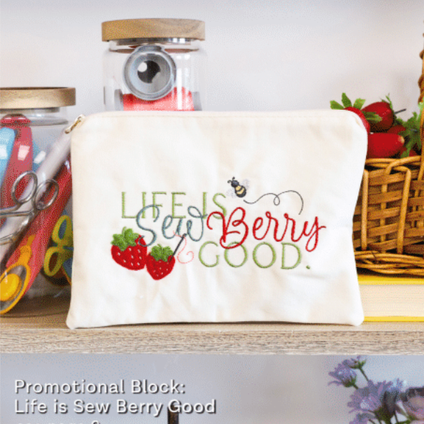 An off-white canvas zippered pouch with embroidered text reading 'Life is Sew Berry Good' and illustrations of strawberries and a bee, displayed on a wooden shelf with sewing-themed props.