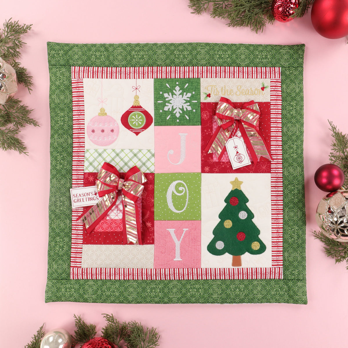 A square quilted Christmas wall hanging or placemat with a green patterned border and a central patchwork design featuring holiday ornaments, a snowflake, the letters J, O, Y, a Christmas tree, and gift boxes with red bows, styled on a pink background with evergreen branches and ornaments.
