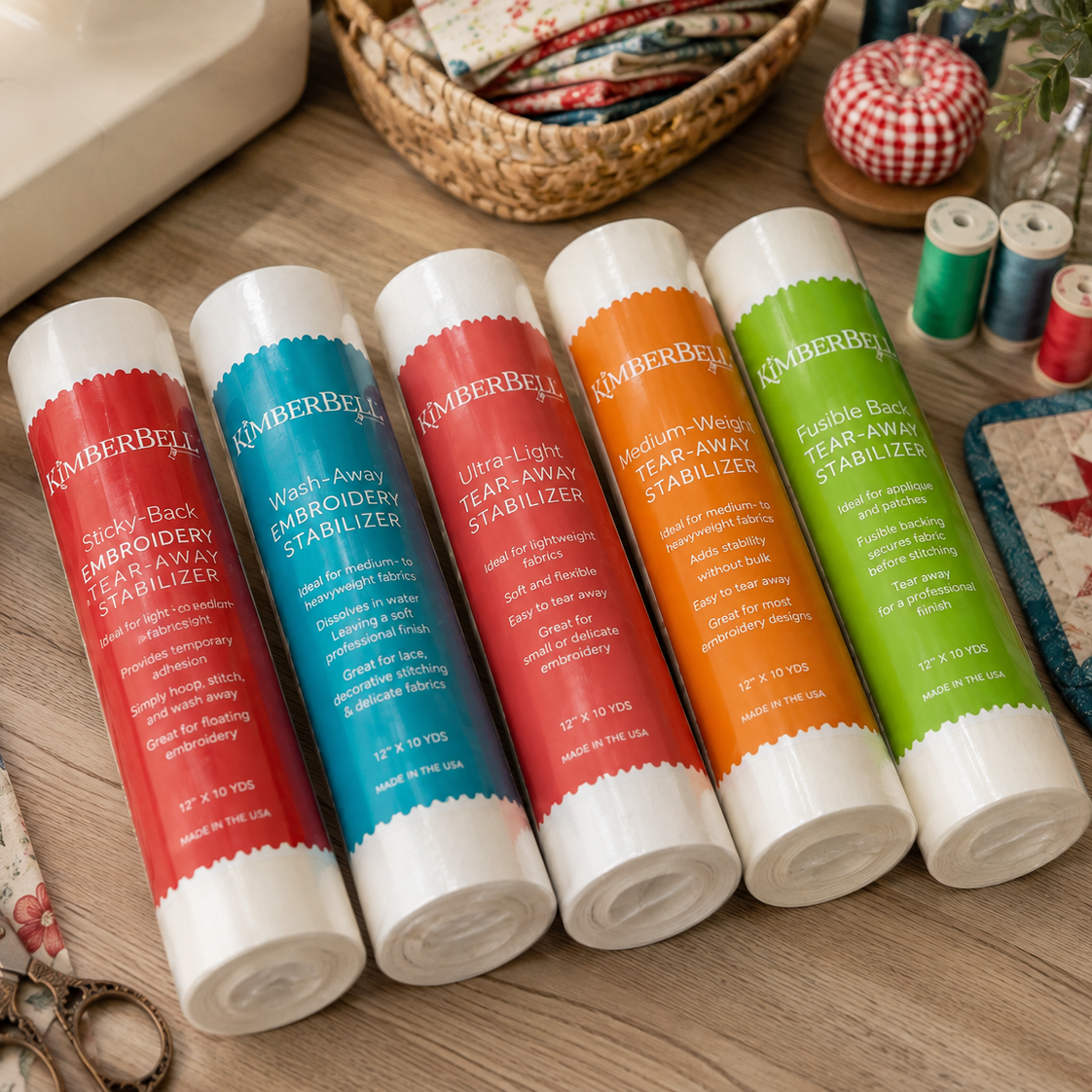Five Kimberly-Clark Sewing Wax Stabilizers in assorted colors (red, teal, coral, orange, and green) arranged on a wooden table surrounded by sewing and crafting props.