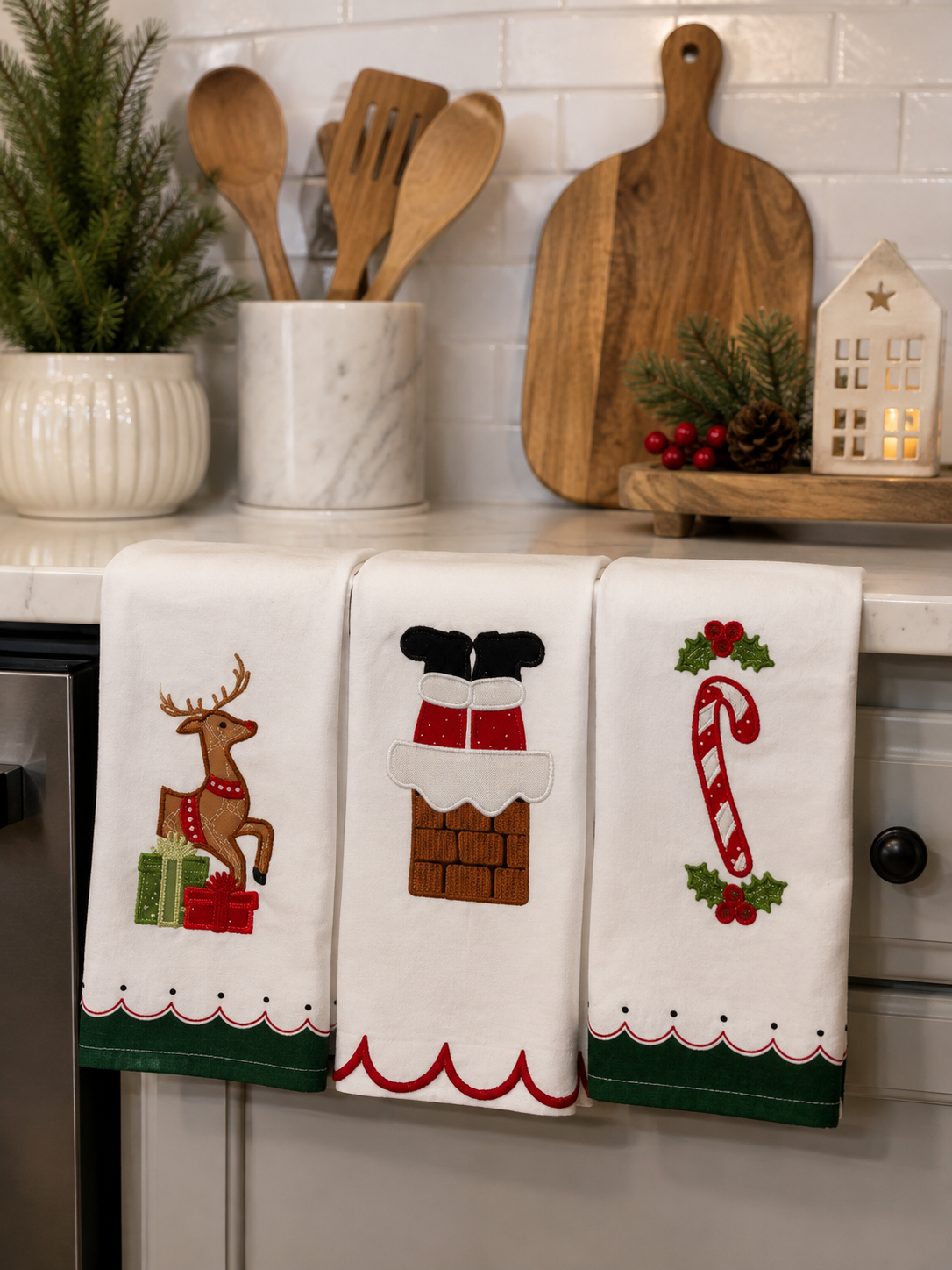 Three white kitchen towels with Christmas-themed embroidery hanging on a countertop. The left towel shows a reindeer with green and red gift boxes, the middle towel shows Santa's boots and chimney, and the right towel shows a candy cane with holly.
