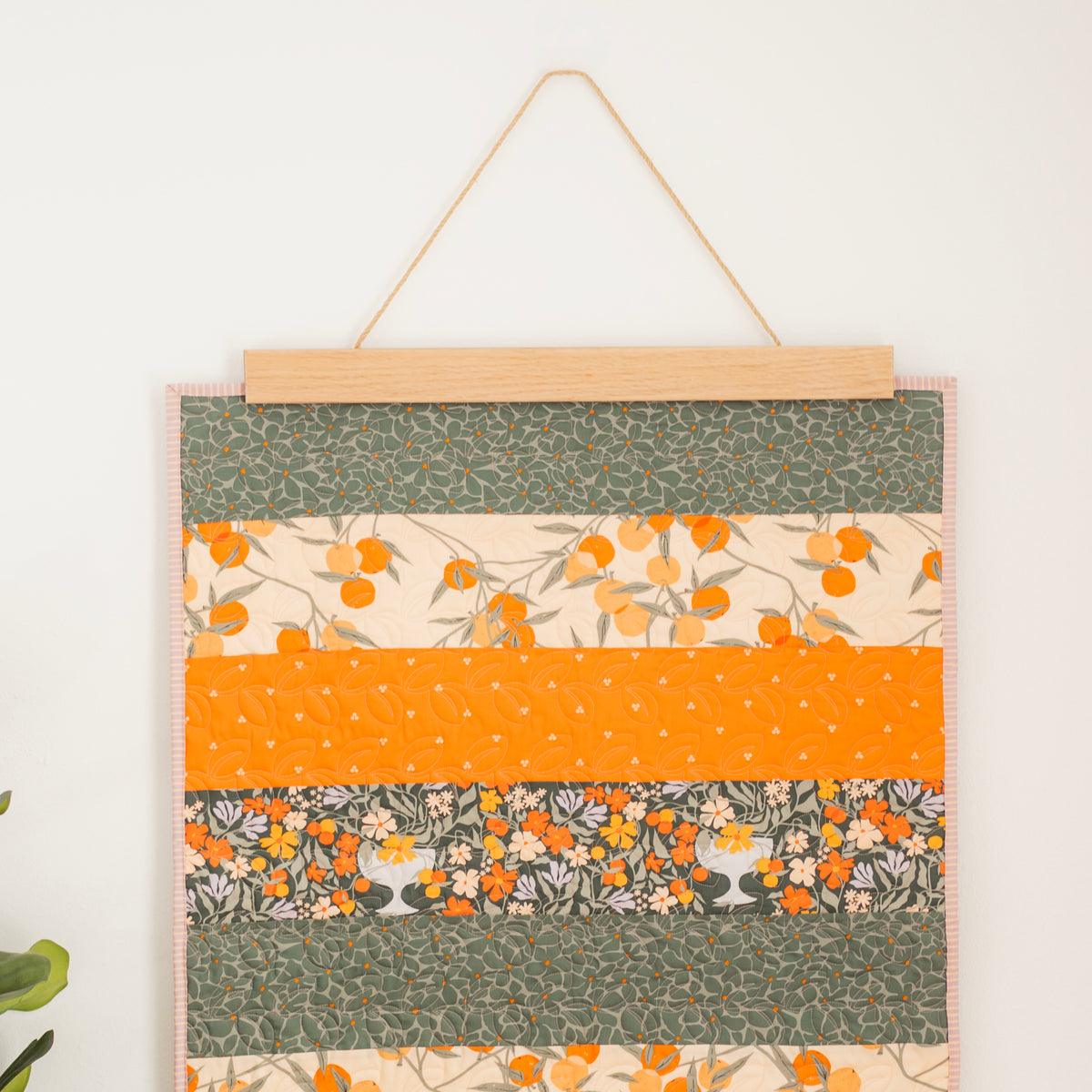 Fabric panel with floral and polka dot patterns in green, orange, and beige hanging on a white wall. 24" wall hanger