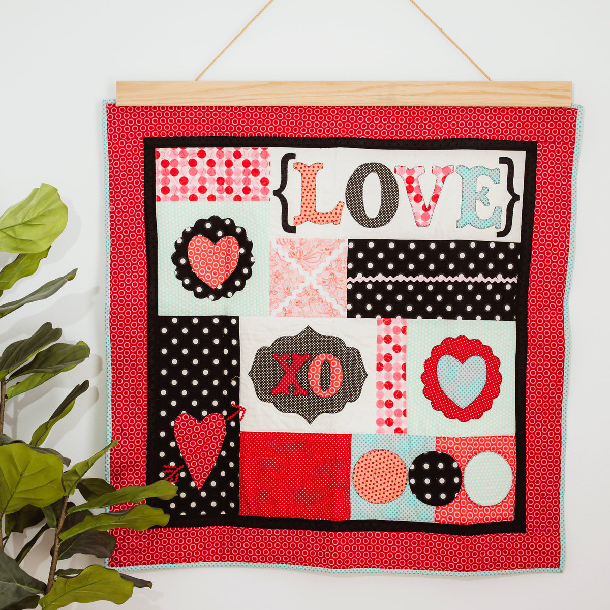 Decorative wall hanging with 'LOVE' and heart designs on a red background, hanging on a white wall with a plant to the left. 36" wall hanger